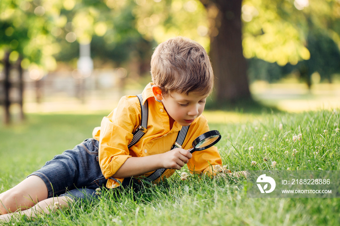 Cute adorable Caucasian boy looking at plants grass flowers in park through magnifying glass. Kid with loupe studying learning nature outside. Child natural science education concept.