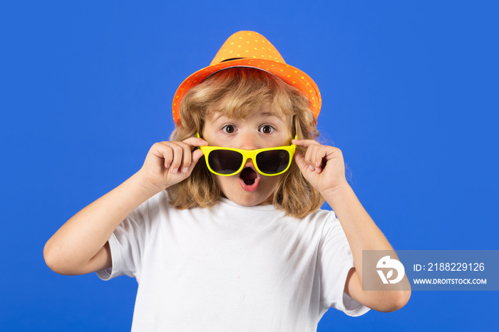 Fashion portrait of kid in summer hat, t-shirt and sunglasses on blue studio isolated background. Excited kid boy on studio isolated background. Surprised face, amazed emotions of child.