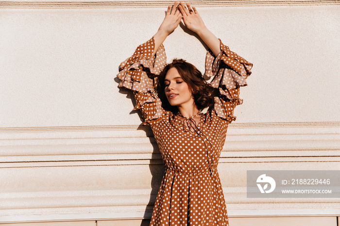 Chilling girl in vintage brown attire posing in front of wall. Ecstatic european young woman standing with hands up.