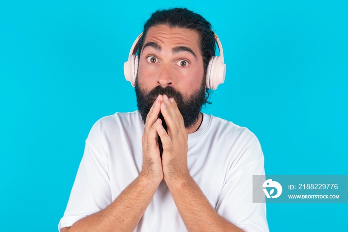 Shocked young bearded man wearing white T-shirt over blue studio background stares fearful at camera keeps mouth widely opened wears wireless stereo headphones on ears