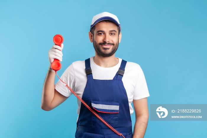 Housing maintenance call centre. Friendly handyman in overalls, safety gloves, holding retro phone handset, answering customer call. Plumber and repair services. indoor studio shot, isolated