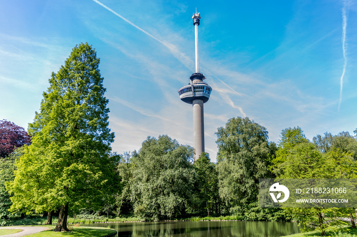 Landscape of a public park with green foliage trees surrounding a pond with the Euromast tower in the background, sunny day with a blue sky and some white clouds in Rotterdam, Netherlands