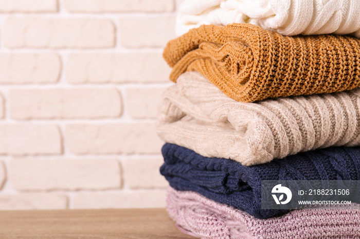 Bunch of knitted pastel color sweaters with different knitting patterns perfectly folded in stack on brown wooden table, white brick wall background. Fall winter season knitwear. Close up, copy space.