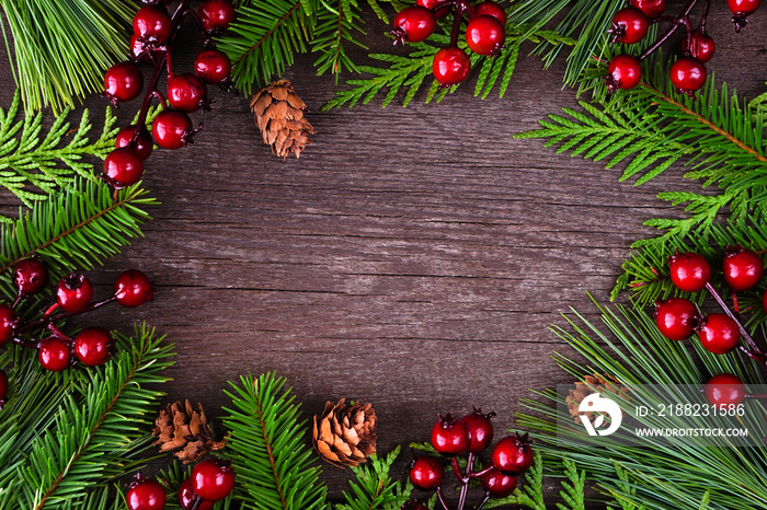 Christmas frame with evergreen branches, red berries and pine cones. Top down view on a dark wood background with copy space.