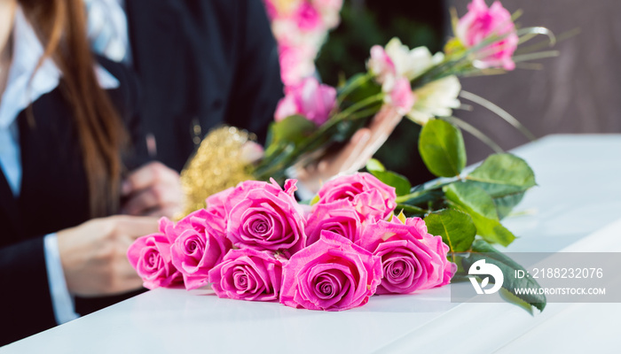 Mourning man and woman on funeral with pink rose standing at casket or coffin, a flower arrangement is in the foreground