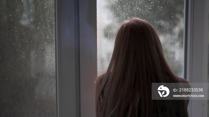 An unrecognizable, bored, lonely girl sits at home due to bad rainy weather and looks out the window at the falling raindrops. Wait for good weather. Back view.