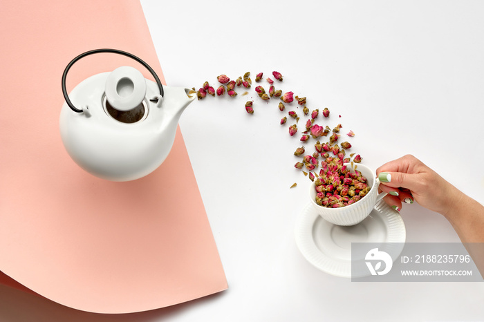 Dry rose flower herbal tea. Surreal flat lay with dried rose flowers pouring from white ceramic tea pot into cup in hand. Herbal tea flower background in pink and off white. Split paper background.