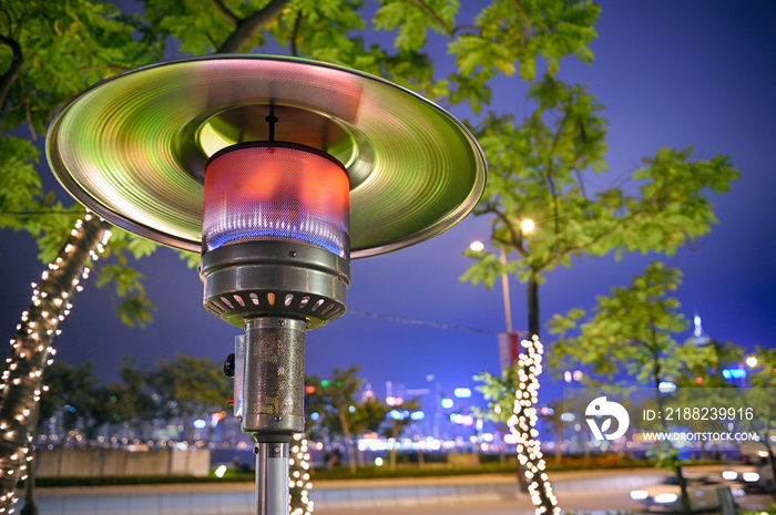 Close up at radiant outdoor gas heater at night in the street restaurant. Green-orange toned reflections. City lights in background.