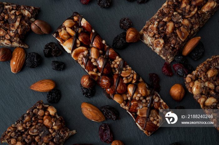 Healthy snacks, fitness lifestyle and high fiber diet concept with granola energy bars surrounded by dried fruits, hazelnuts and almonds on a black stone with dramatic light