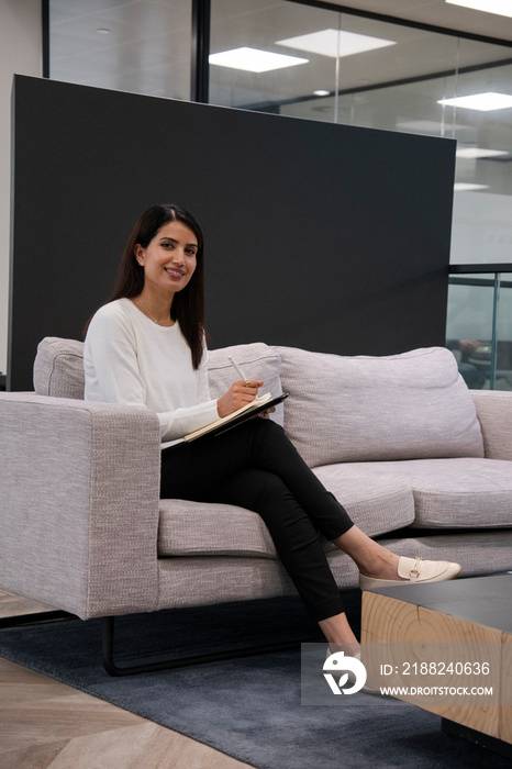 Portrait of woman sitting on sofa and writing in notepad