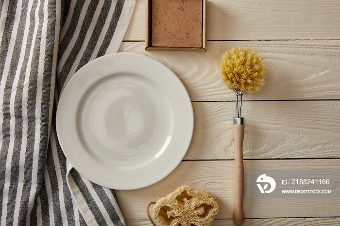 top view of empty white plate, striped towel and different cleaning items on white wooden surface, zero waste concept