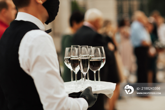 Waiter in black mask and gloves hold tray with glasses of champagne. Waiter holding a serving tray full of drinks in champagne glasses. Wearing masks during coronavirus.