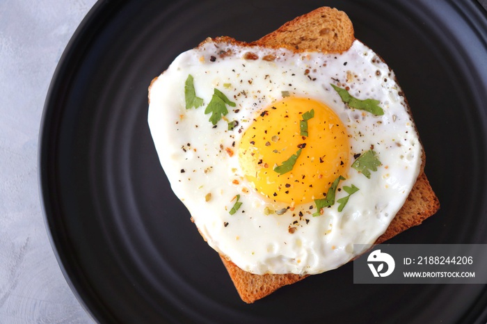 Fried Egg on Whole wheat Toast with salt and pepper for classic Breakfast. sunny side up egg with brown bread on black plate over wooden table, top view, copy space.