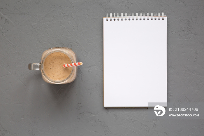 Homemade smoothie with coffee, oat and banana in a glass jar mug, blank notepad on a gray surface, top view. Overhead, from above.