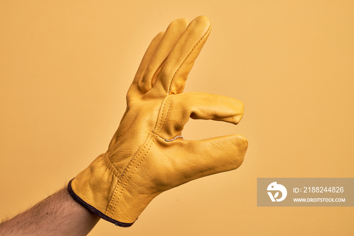 Hand of caucasian young man with gardener glove over isolated yellow background picking and taking invisible thing, holding object with fingers showing space