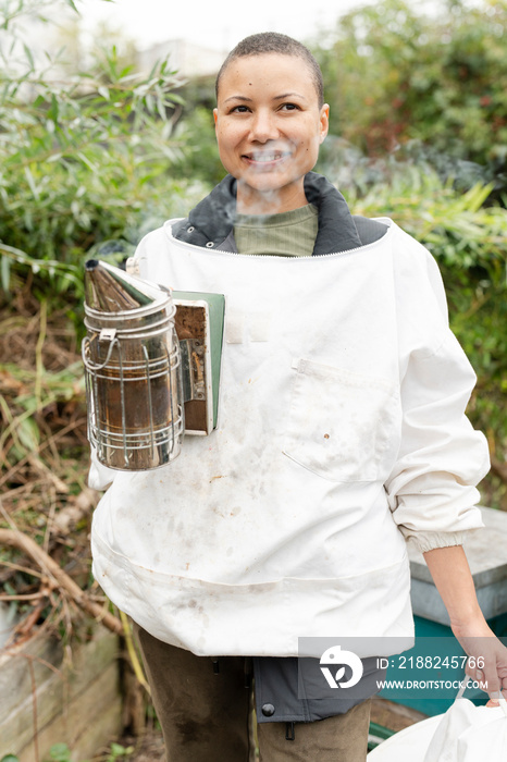 Portrait of smiling female beekeeper holding smoker in urban garden