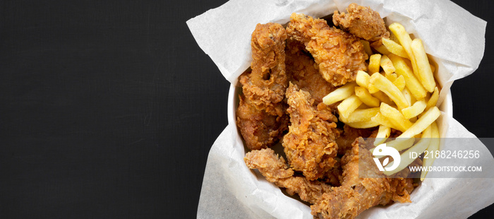 Fried chicken legs, spicy wings, French fries and chicken fingers in paper box over black background, top view. Flat lay, overhead, from above. Copy space.