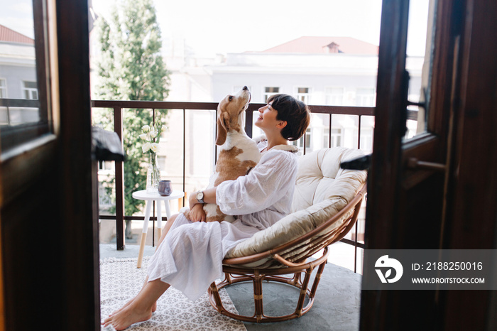 Relaxed young woman in white cozy bathrobe sitting with beagle dog on balcony in summer morning. Pretty brunette girl in wristwatch holding puppy on knees and smiling.