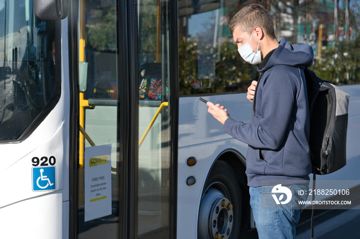 Young caucasian man wearing face mask and looking at mobile while taking the bus during pandemic covid-19