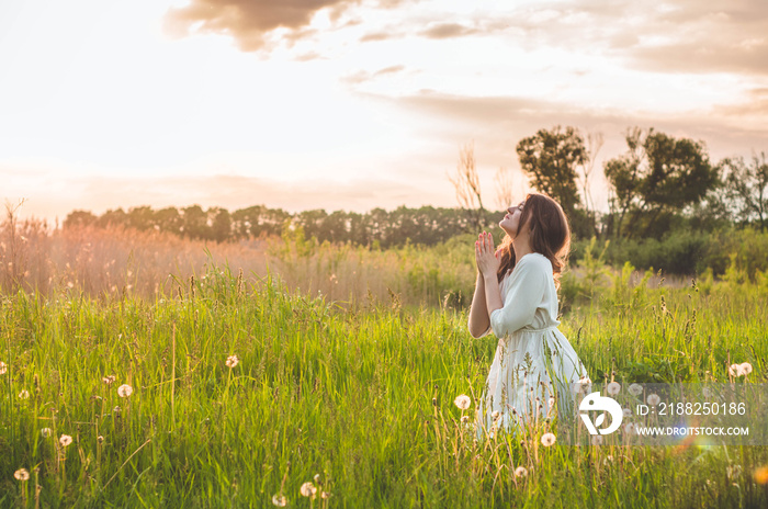 Girl closed her eyes, praying in a field during beautiful sunset. Hands folded in prayer concept for faith, spirituality and religion. Peace, hope, dreams concept