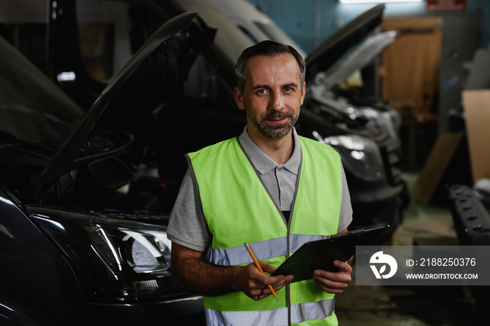 Waist up portrait of mature male worker looking at camera while inspecting vehicles at car factory quality control