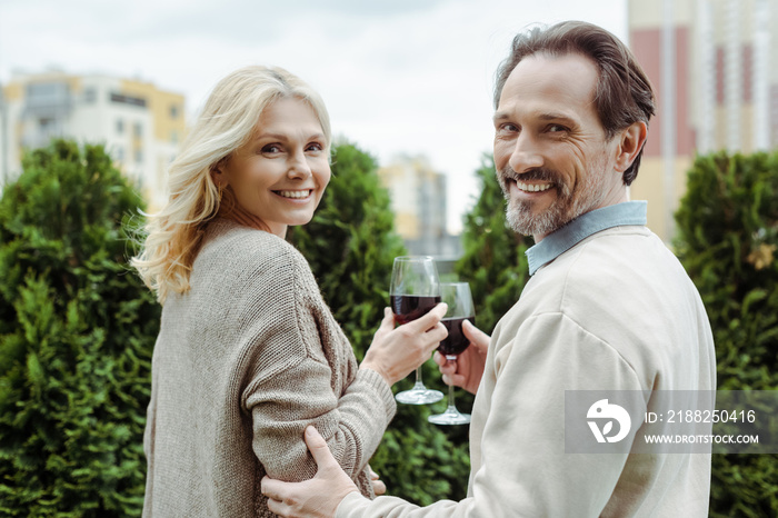 Side view of mature couple smiling at camera while holding glasses of wine on urban street