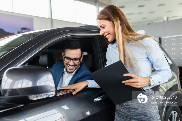 Saleswoman at the dealership showroom talking with customer and helping him to choose a new car for himself.