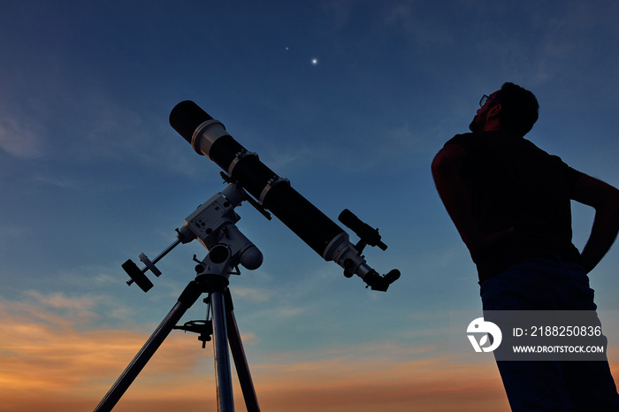 Silhouette of a man, telescope and countryside under the starry skies.