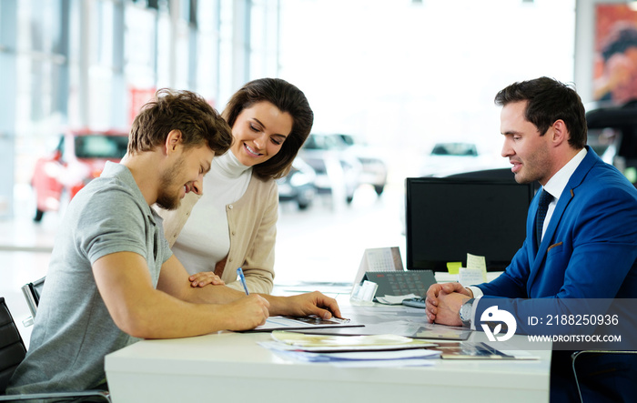 Beautiful young couple signs documents at dealership showroom