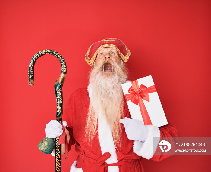 Old senior man with grey hair and long beard wearing saint nicholas costume holding present angry and mad screaming frustrated and furious, shouting with anger looking up.