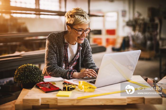 Cheerful smiling beautiful middle-aged female engineer looking on a laptop on the desk in the sunny workshop.