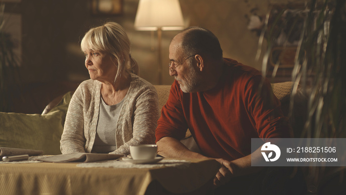 Irritated aged husband blaming upset wife while sitting on sofa at home together