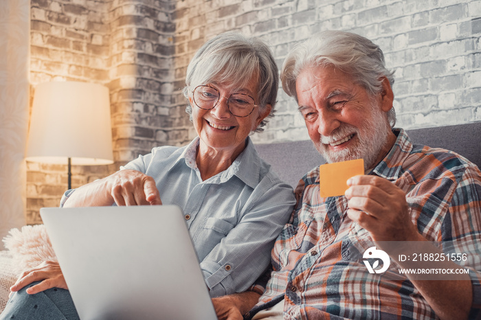 Portrait of couple of cute two happy and excited seniors buying and spending money shopping online with laptop on the sofa at home. Using credit card together buying on internet.