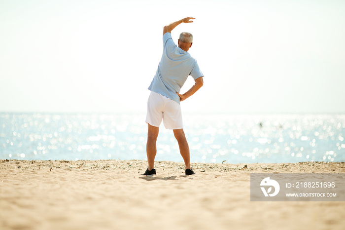 Mature man in activewear standing on sand in front of waterside and side-bending in the morning