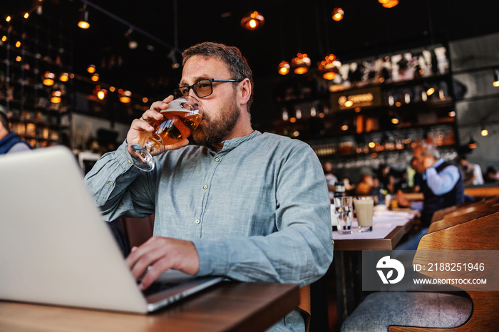 Middle aged bearded freelancer with eyeglasses sitting in a bar, drinking fresh cold light beer and typing on laptop.