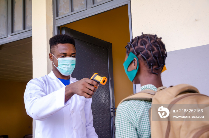 a young handsome african class teacher holding as thermometer to scan the temperature of his student before entering the classroom