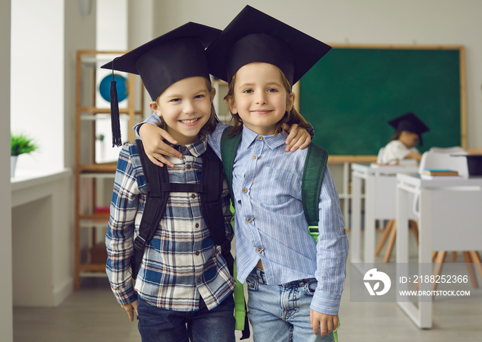 Portrait of two cheerful little school boys in mortar boards who are best friends in class. Children in shirts, jeans and graduation hats hug and smile looking at the camera. Graduation concept.