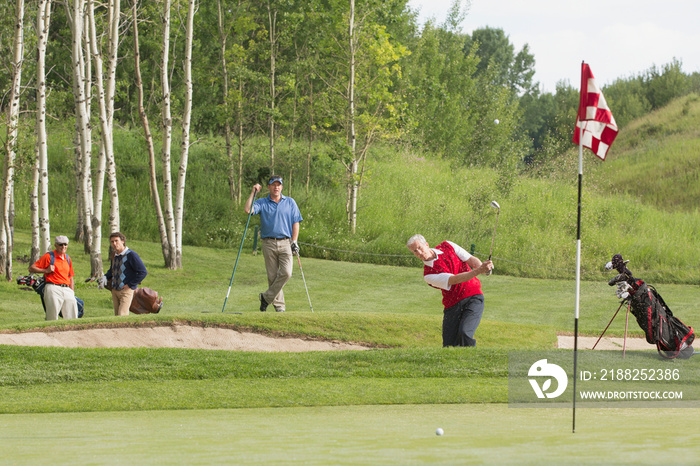 Female golfer chipping out of sand trap as other golfers watch