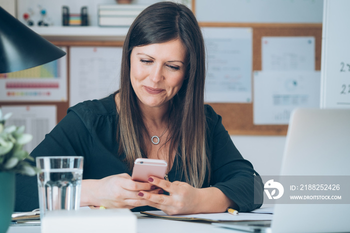 Close up photo of smiling female teacher sitting at desk and holding mobile phone. Happy professor or student having break with online class and using her smartphone to send text message or video call