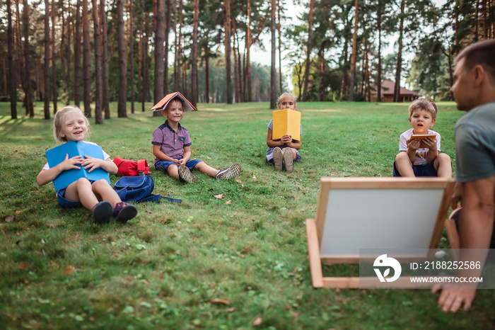 Back to school. Kindergarten and elementary scholars sitting with teacher on grass at open-air class