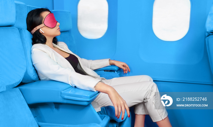 Side view of a young businesswoman in white suit sleeping during flight on the airplane at the first class. Female in sleeping mask enjoying her comfortable flight while sitting in the airplane cabin.