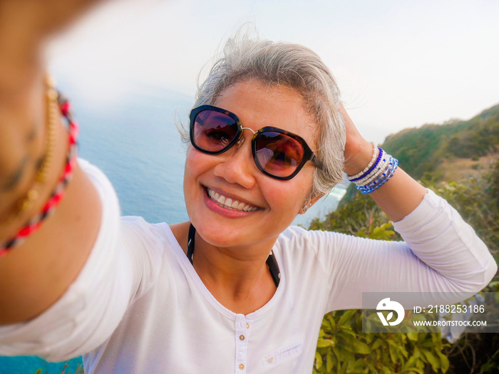 middle age 50s happy and cheerful Asian woman with grey hair taking selfie with mobile phone at beautiful tropical beach island smiling at cliff viewpoint enjoying Summer