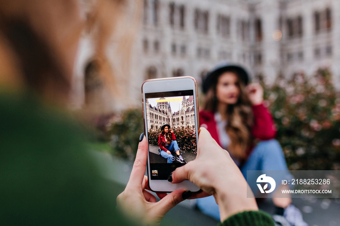 Girl in vintage jeans posing near historical place for her sister. Female photographer using phone for taking picture of friend.