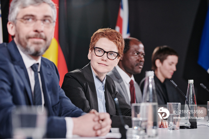 Young red haired woman in eyeglasses sitting at table among her international colleagues during press conference