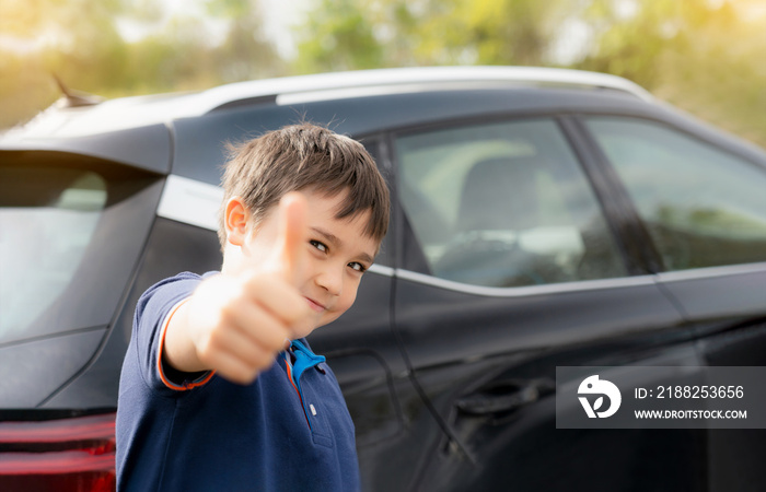 Happy young boy showing thumb up while stnading next to the car, School kid with smiling face standing at the carpark in sunny day Spring or Summer, Positive child going to school.