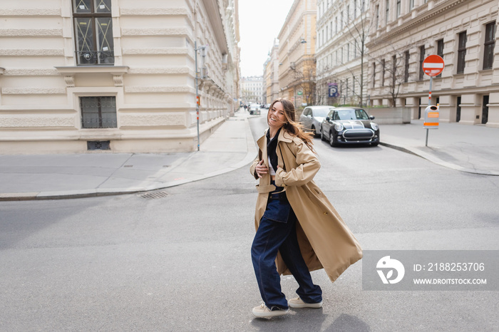 full length of cheerful young woman in beige trench coat walking on european street.