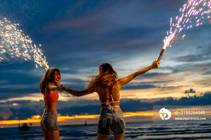 Young Asian woman having fun dancing and playing sparklers together on tropical island beach in summer night. Happy female friends enjoy outdoor lifestyle nightlife on holiday travel vacation trip