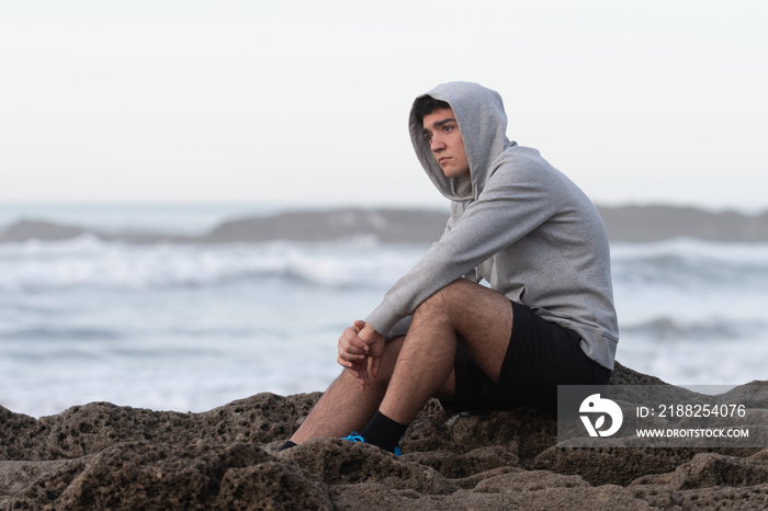 Worried hispanic teenager sitting on rocks beside the sea. Anxiety and depression on adolescence concept.