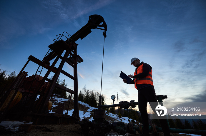 Low angle of petroleum engineer in work vest looking at oil pump rocker-machine and writing on clipboard. Oil worker checking work of balanced beam petroleum pump jack under beautiful evening sky.