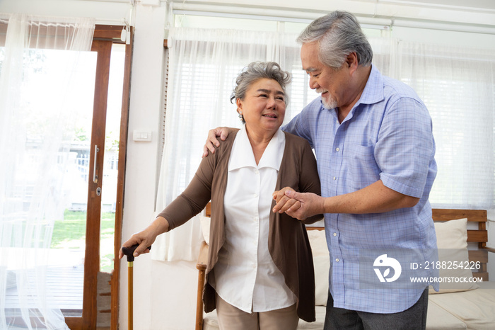 Happy senior asian man helps older wife with a cane walk through their house by holding her hand